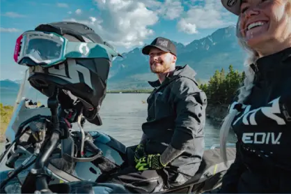 A man and woman smiling while seated on a CFMOTO IBEX motorcycle, with a scenic landscape in the background and sunlight behind them.