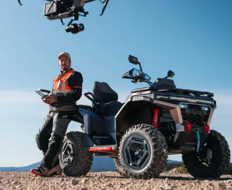 A man wearing an orange work jacket is standing and leaning against the rear of a CFMOTO CFORCE C6 Touring while piloting a drone.